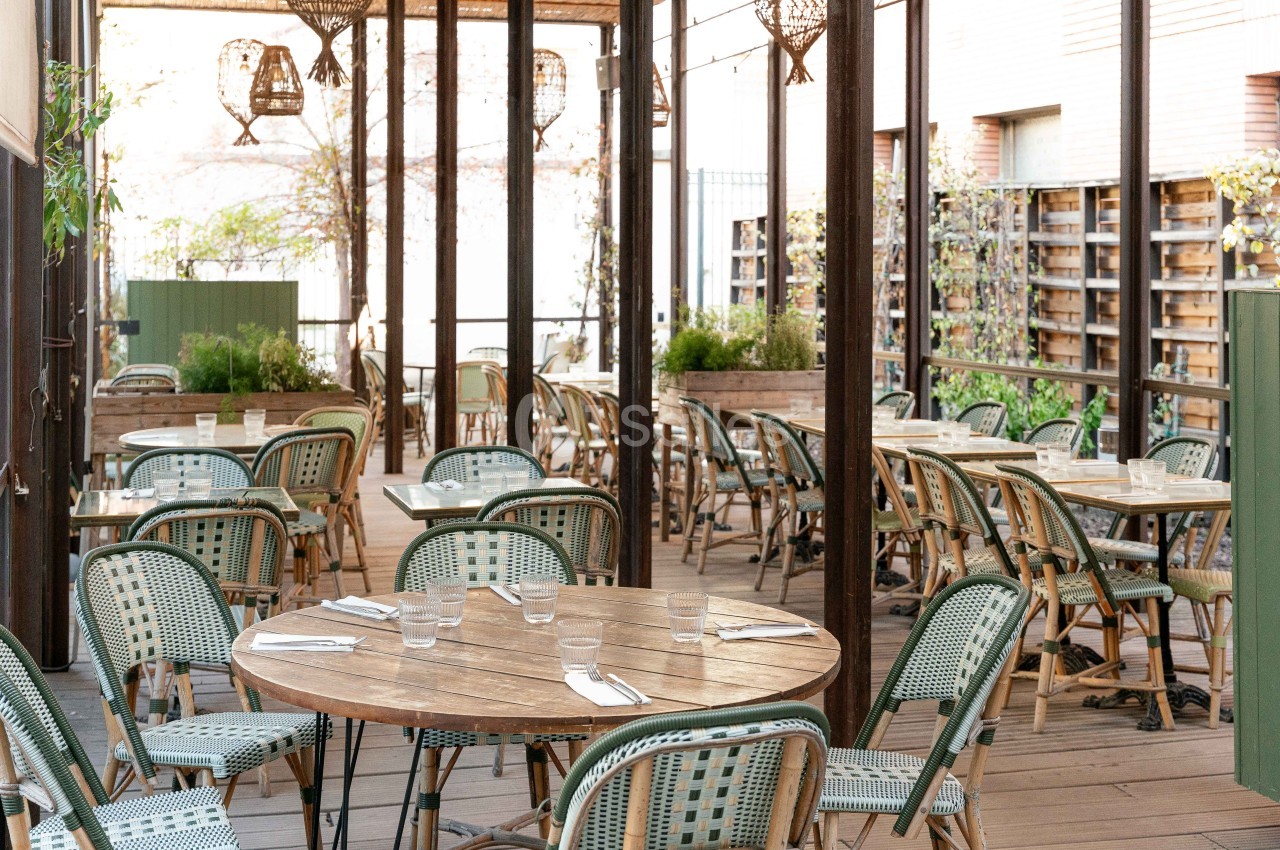 Terrasse couverte avec tables rondes en bois, chaises en rotin vert et blanc, paniers suspendus, vue sur jardin.