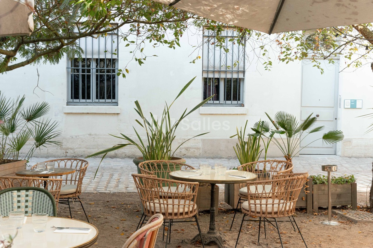 Terrasse ombragée avec chaises en rotin autour de tables rondes, façade blanche et plantes en arrière-plan.