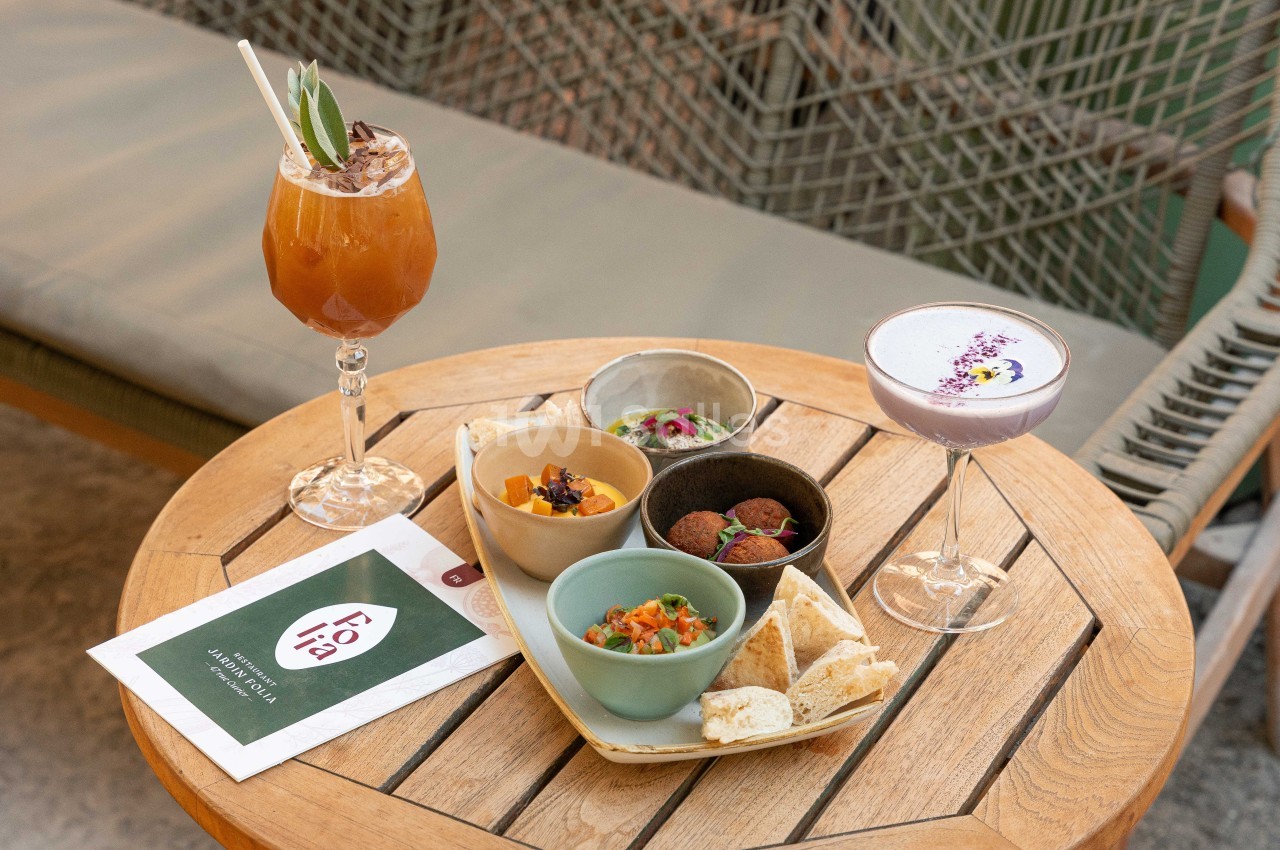 Table ronde en bois dressée en terrasse avec cocktails, assiettes de mets et menu, chaises en rotin à l'arrière.