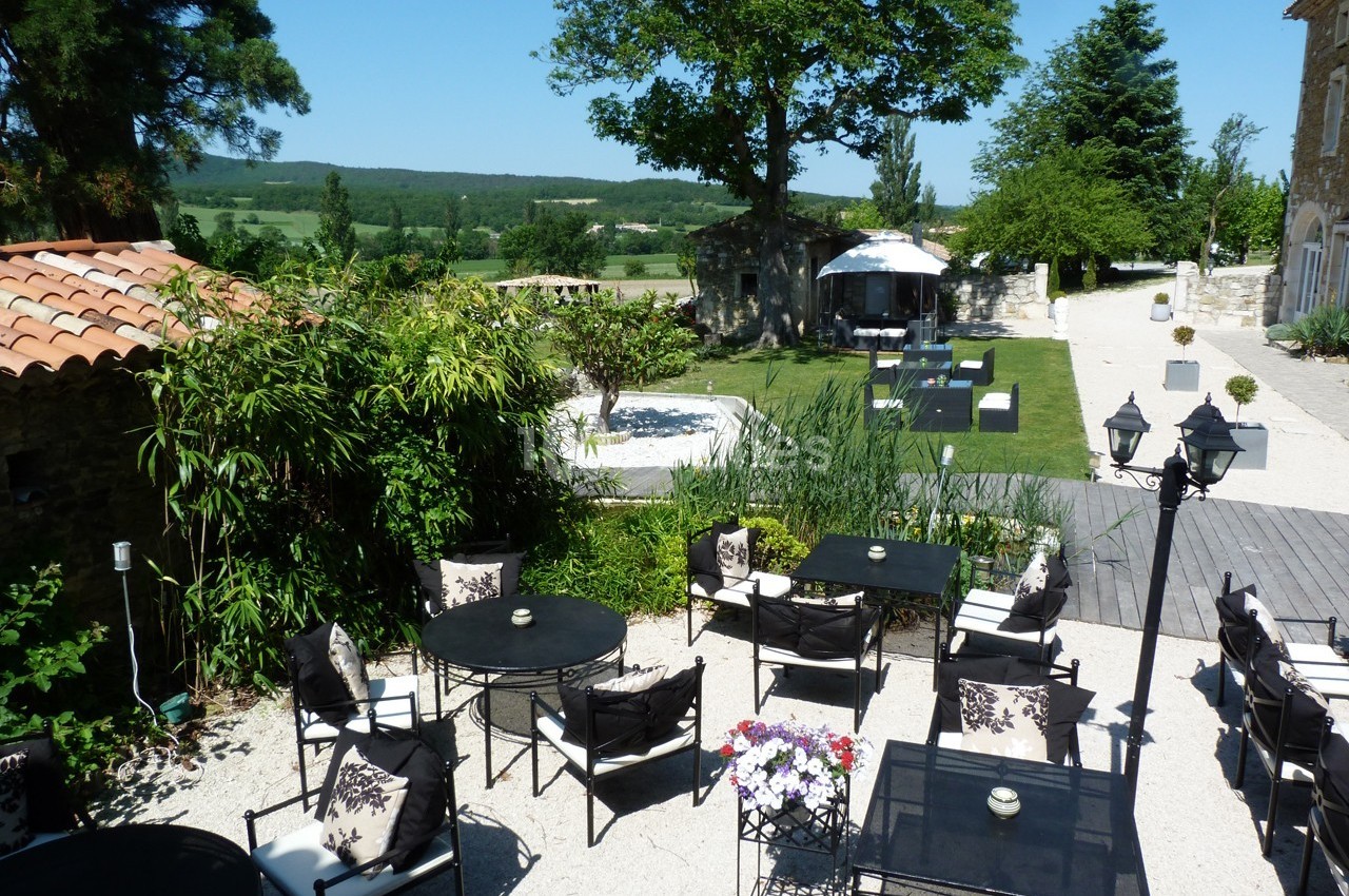 Terrasse extérieure avec tables et chaises, jardin paysager, pelouse, arbres et vue sur la campagne environnante.