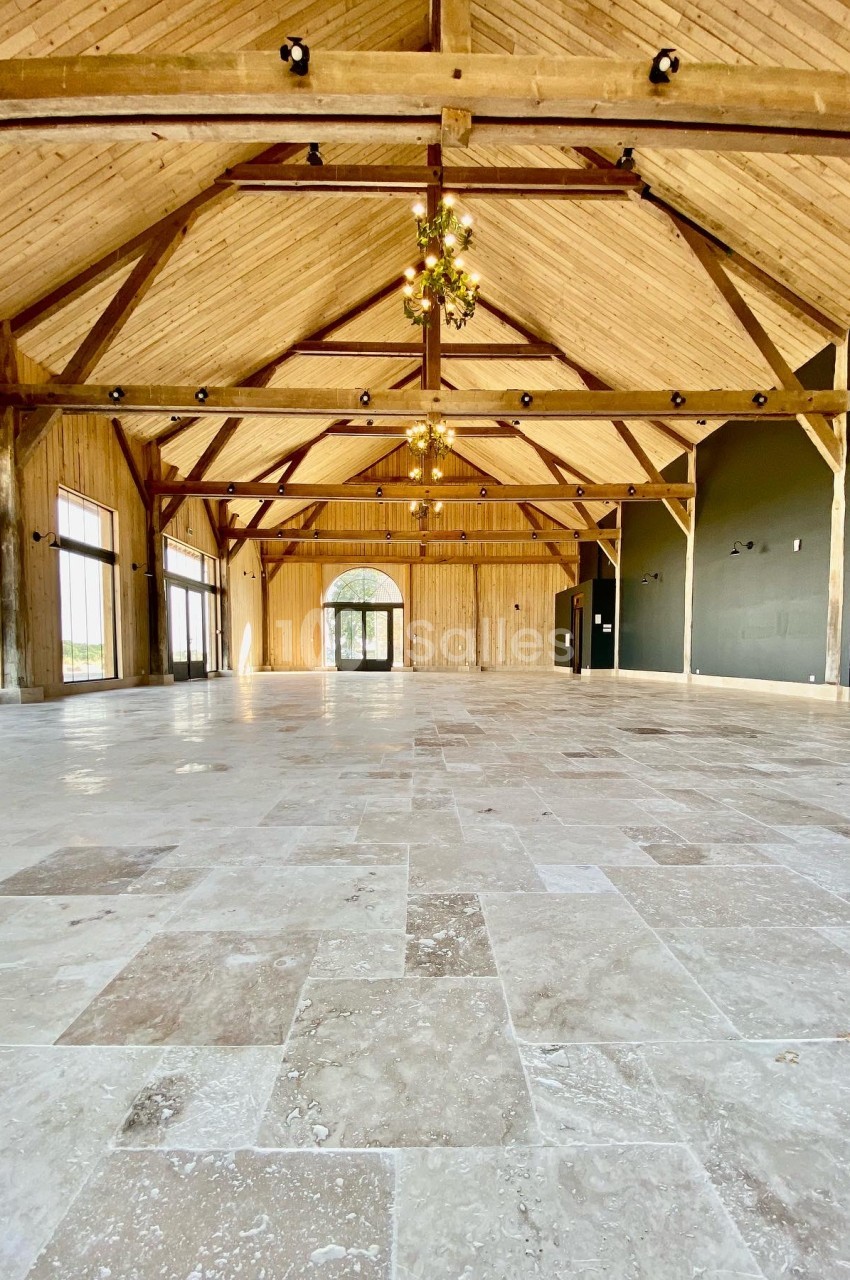 Salle avec charpente bois apparente, sol dallé, poutres apparentes et lustre central, vide.