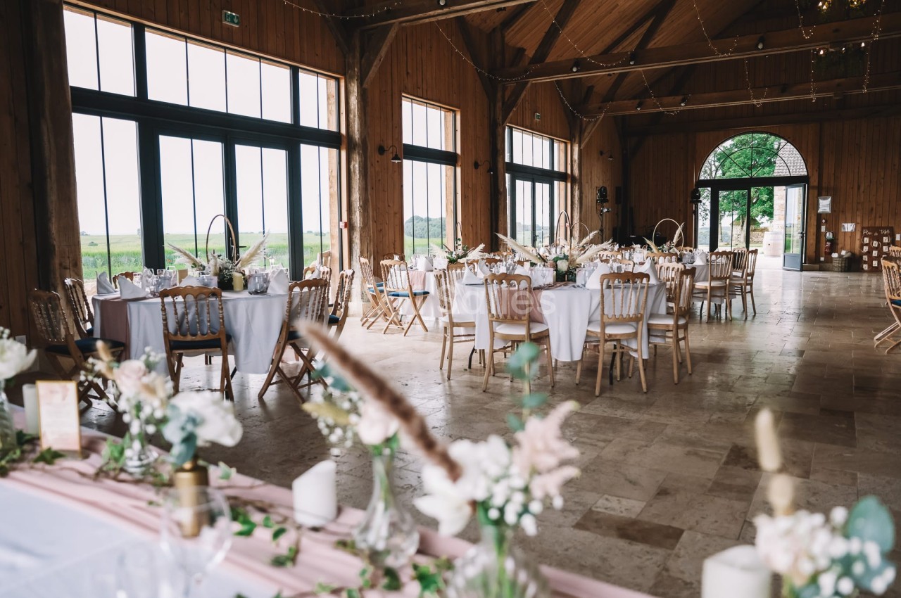 Salle en bois avec poutres apparentes, tables rondes dressées en blanc, chaises bois naturel, grandes baies vitrées sur paysage rural.