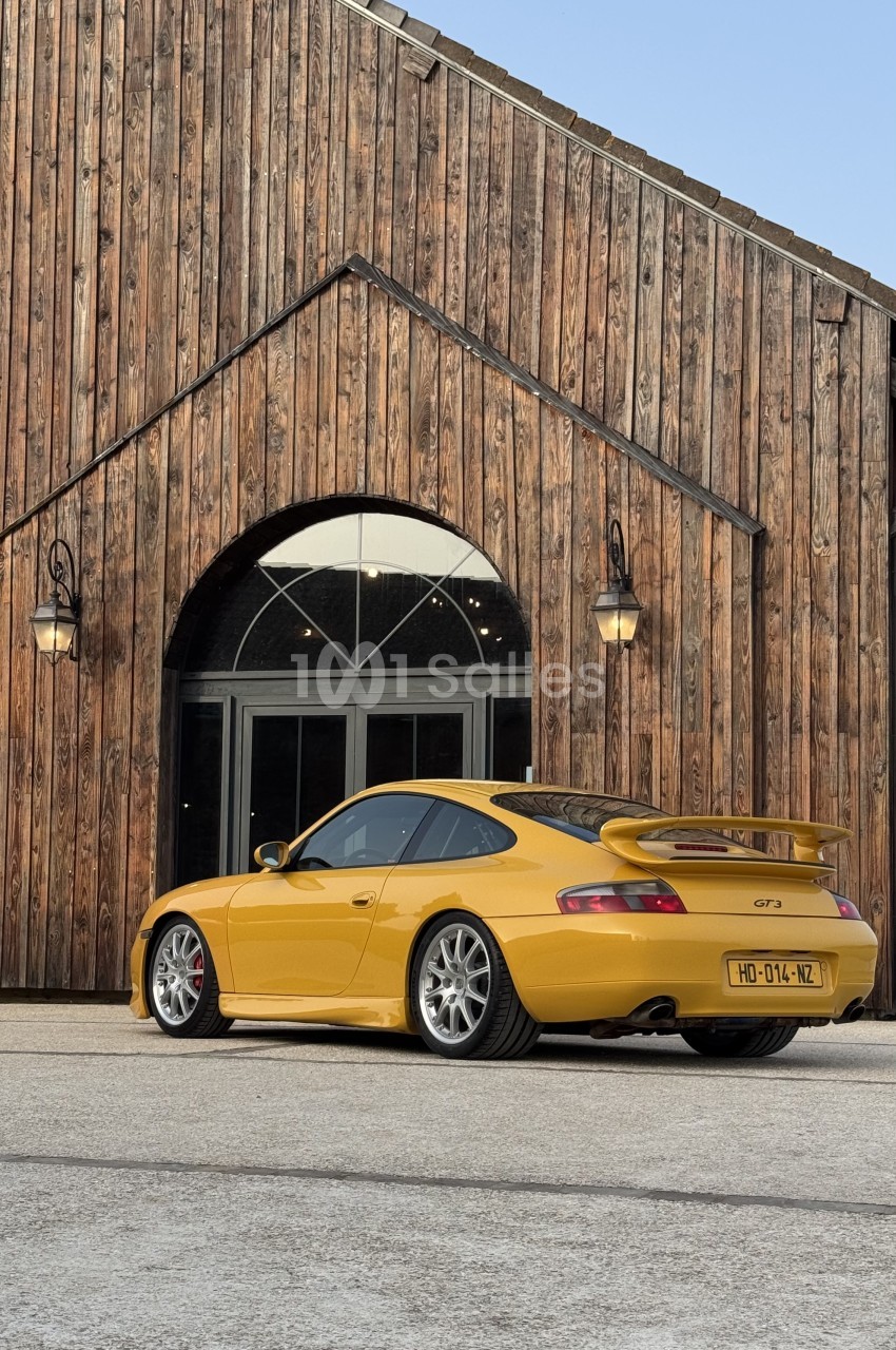 Porsche GT3 jaune devant grange en bois avec porte cintrée et lanternes noires.