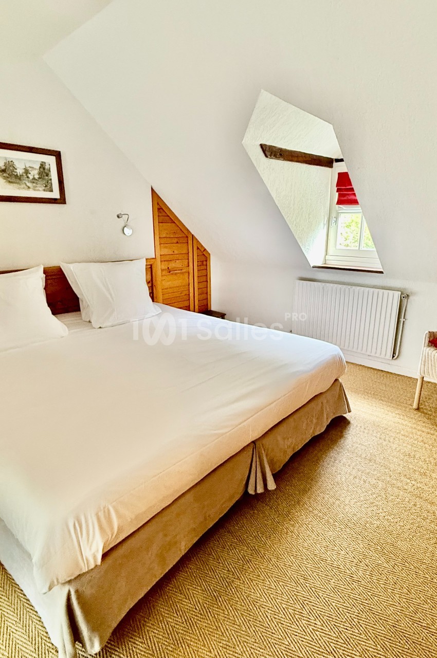 Chambre mansardée avec un grand lit recouvert de linge blanc, une tête de lit en bois sous les combles et une fenêtre de toit.