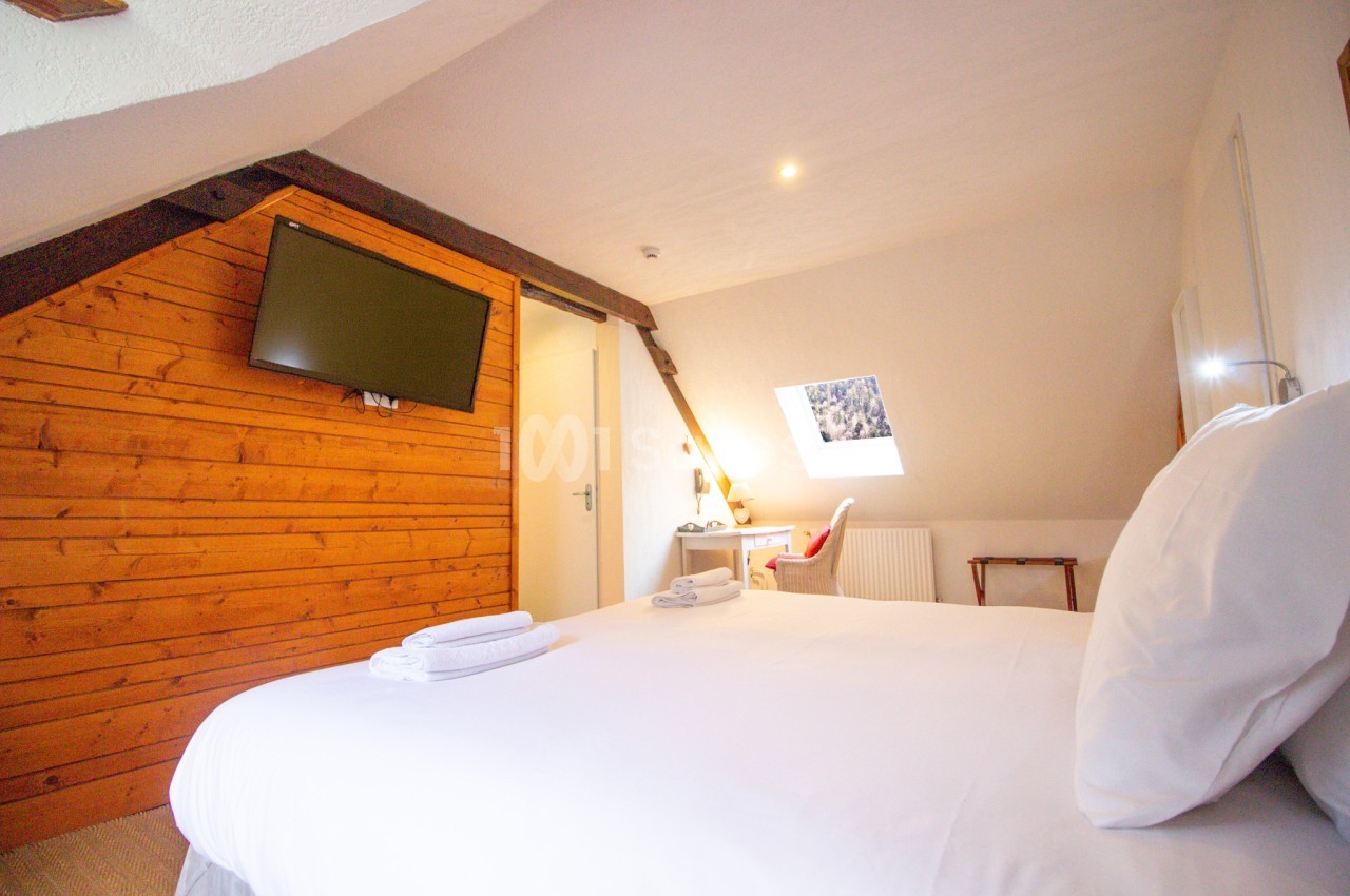 Chambre mansardée avec lit blanc, serviettes pliées sur la couverture, mur en lambris de bois et télévision.