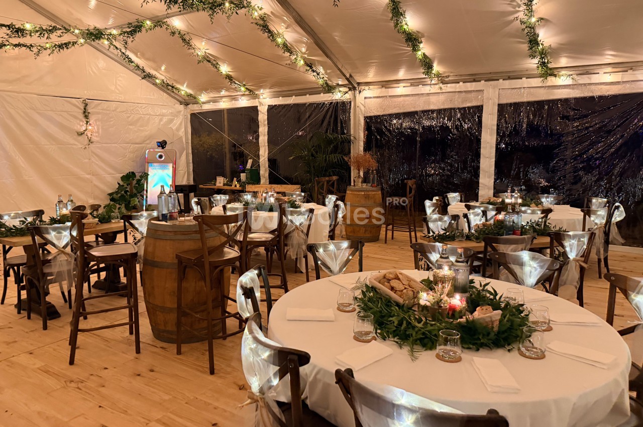 Intérieur d'un chapiteau éclairé la nuit : tables rondes nappées de blanc, chaises en bois avec noeuds de tulle, guirlandes lumineuses et tonneau servant de mange-debout. Let me check constraints: one sentence, under 125 chars? Count: "Intérieur d'un