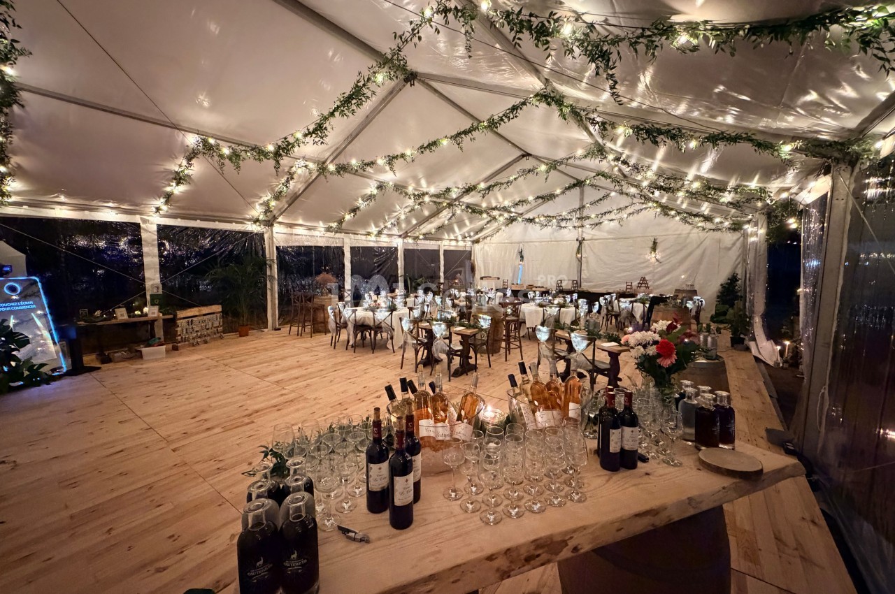 Intérieur d'un chapiteau transparent éclairé de guirlandes lumineuses et de feuillage, avec tables dressées au fond et bar en bois garni de bouteilles et de verres.