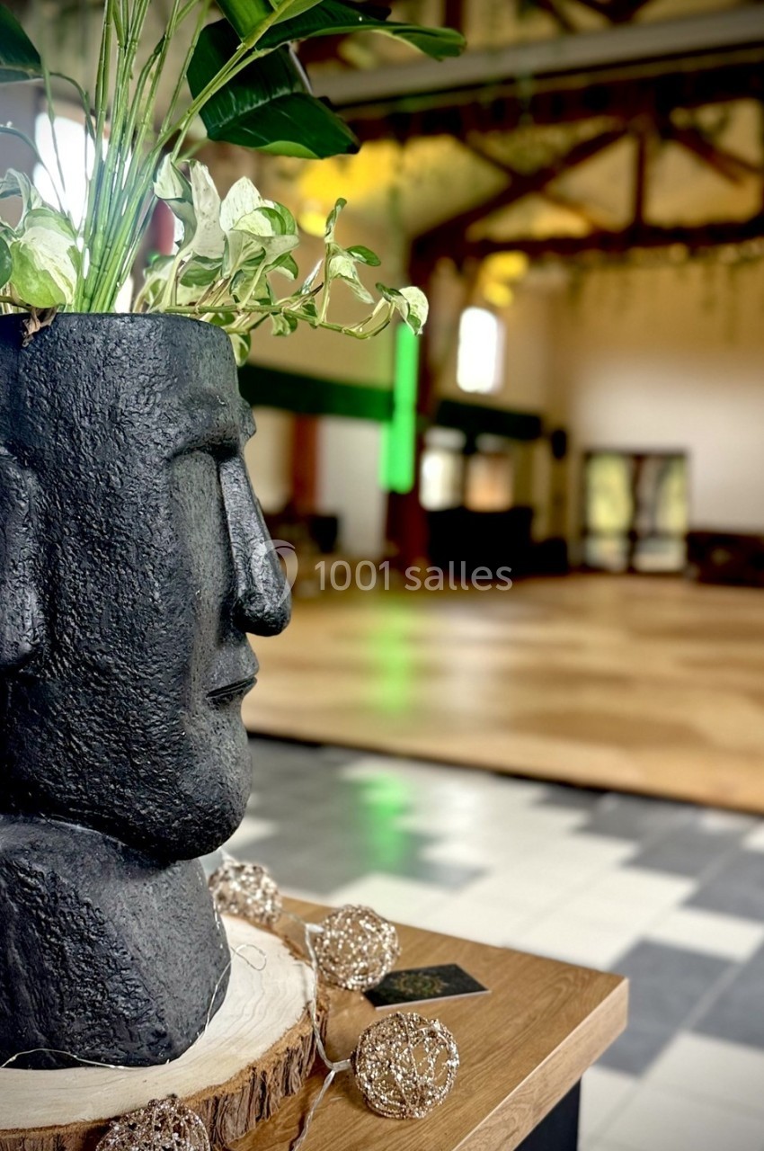 Pot de fleurs en forme de tête sculptée, posé sur une table en bois, dans une grande salle lumineuse.