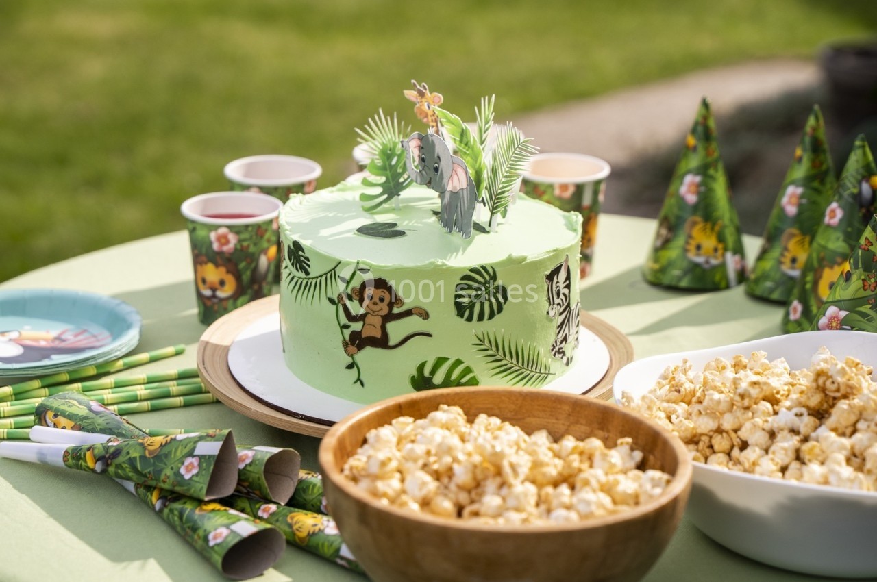 Gâteau décoré sur le thème de la jungle entouré de popcorn, gobelets et chapeaux assortis pour une fête en plein air.