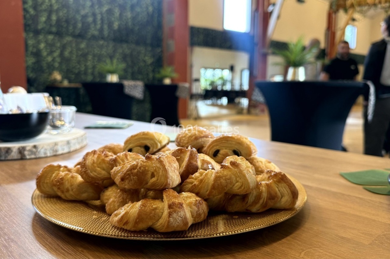 Assortiment de viennoiseries sur un plateau doré, posé sur une table en bois dans une salle décorée de plantes.