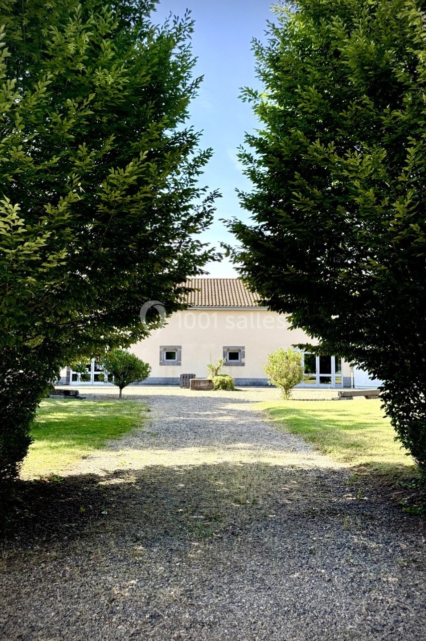 Allée gravillonnée bordée d'arbres menant à une maison beige avec toit en tuiles, entourée de verdure.