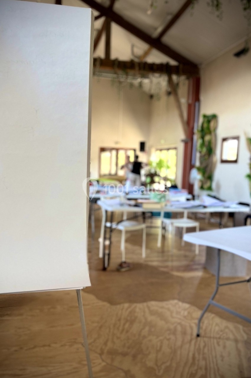 Salle lumineuse avec des tables disposées pour un atelier, vue partielle d'un tableau de conférence au premier plan.