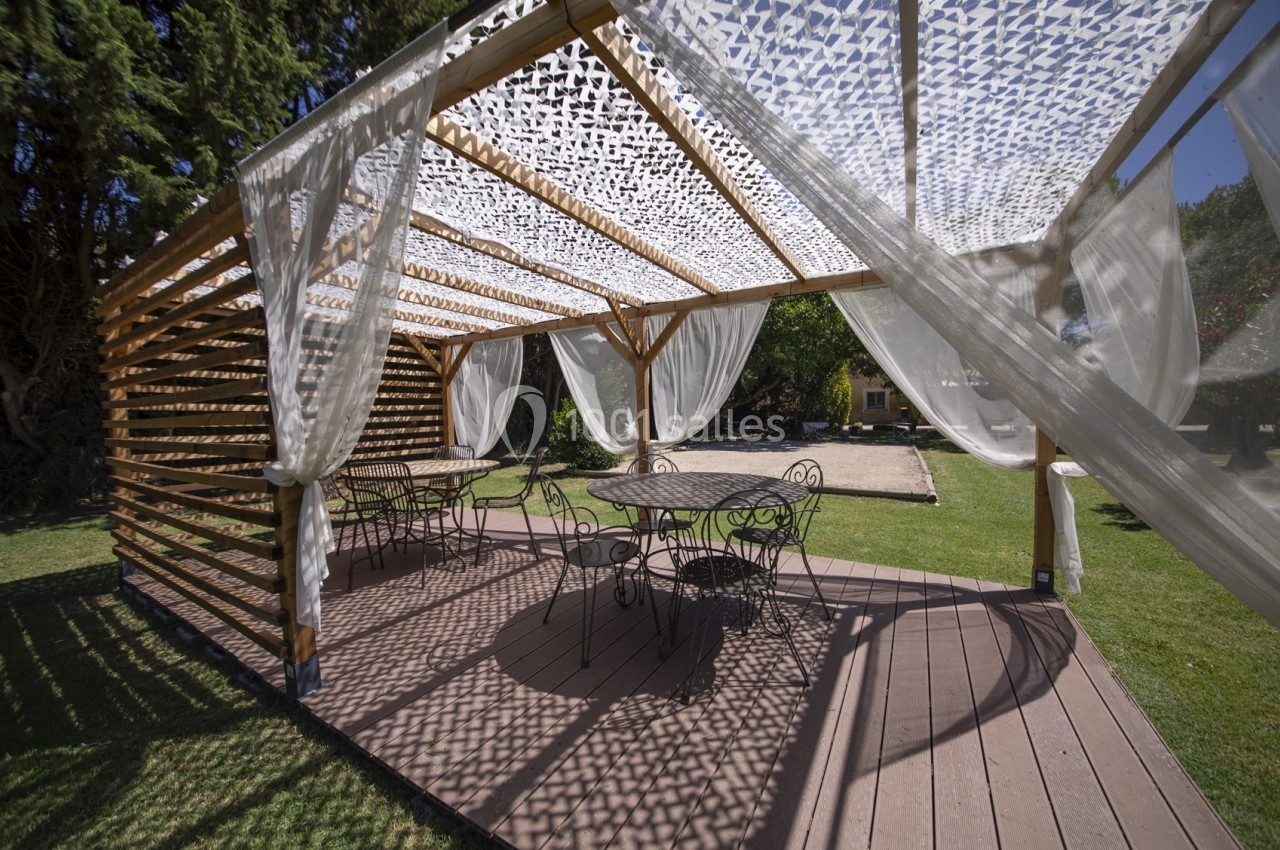 Pergola en bois avec voiles d'ombrage, abritant des tables et chaises en fer forgé sur une terrasse ensoleillée.