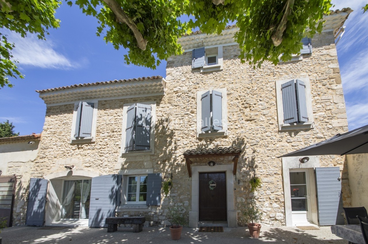Façade en pierre d'une maison provençale avec volets bleus, sous un ciel bleu et des arbres ombrageants.