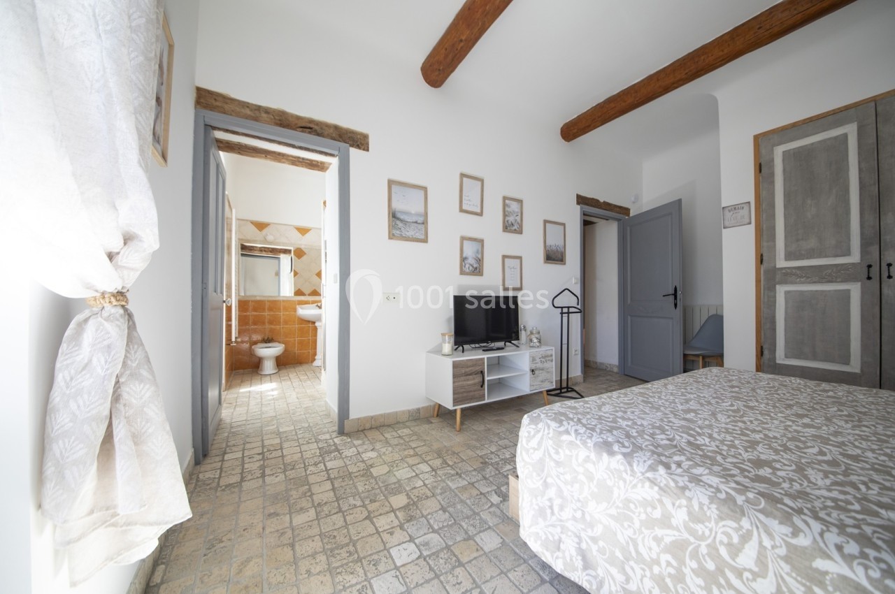Chambre lumineuse avec lit double, télévision, meubles clairs, poutres apparentes et vue sur une salle de bain attenante.