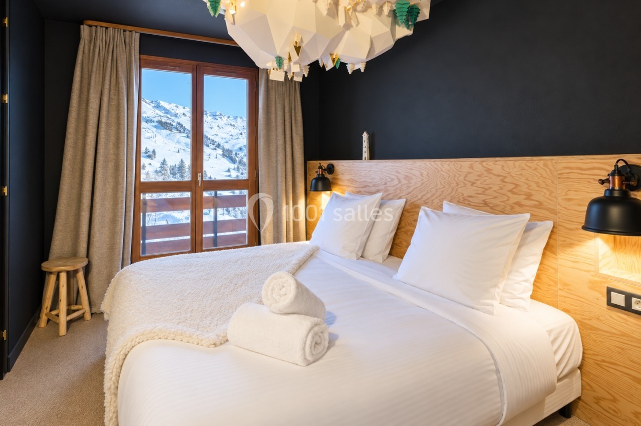 Chambre lumineuse avec lit double, linge blanc, lampe murale et vue sur montagne enneigée depuis une grande fenêtre.