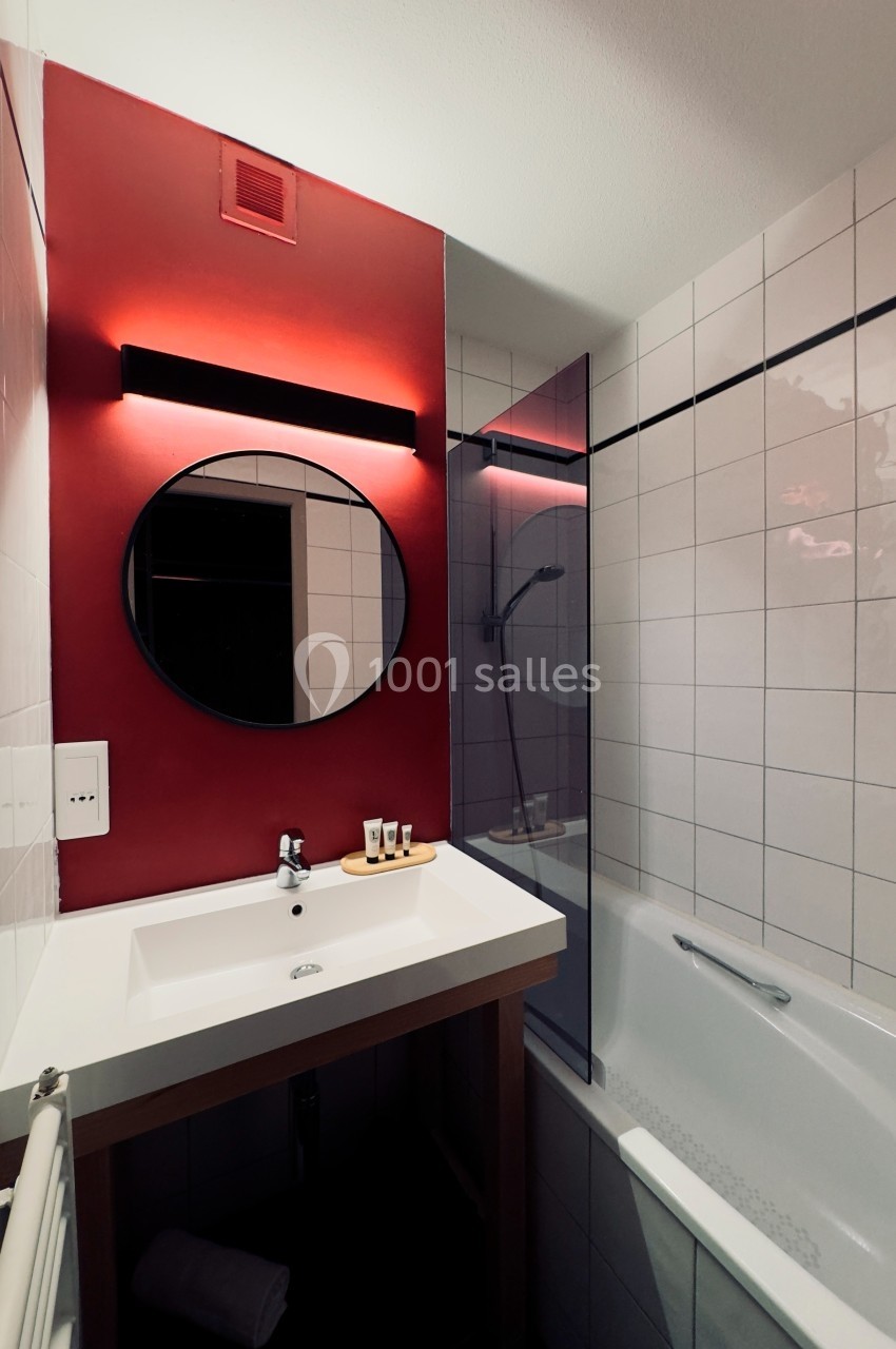 Salle de bain avec lavabo, miroir rond, mur rouge, baignoire et carrelage blanc.
