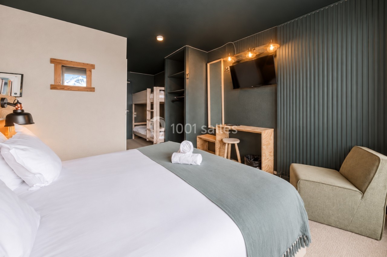 Chambre d'hôtel moderne avec lit double, lits superposés en arrière-plan, télévision murale et éclairage chaleureux.