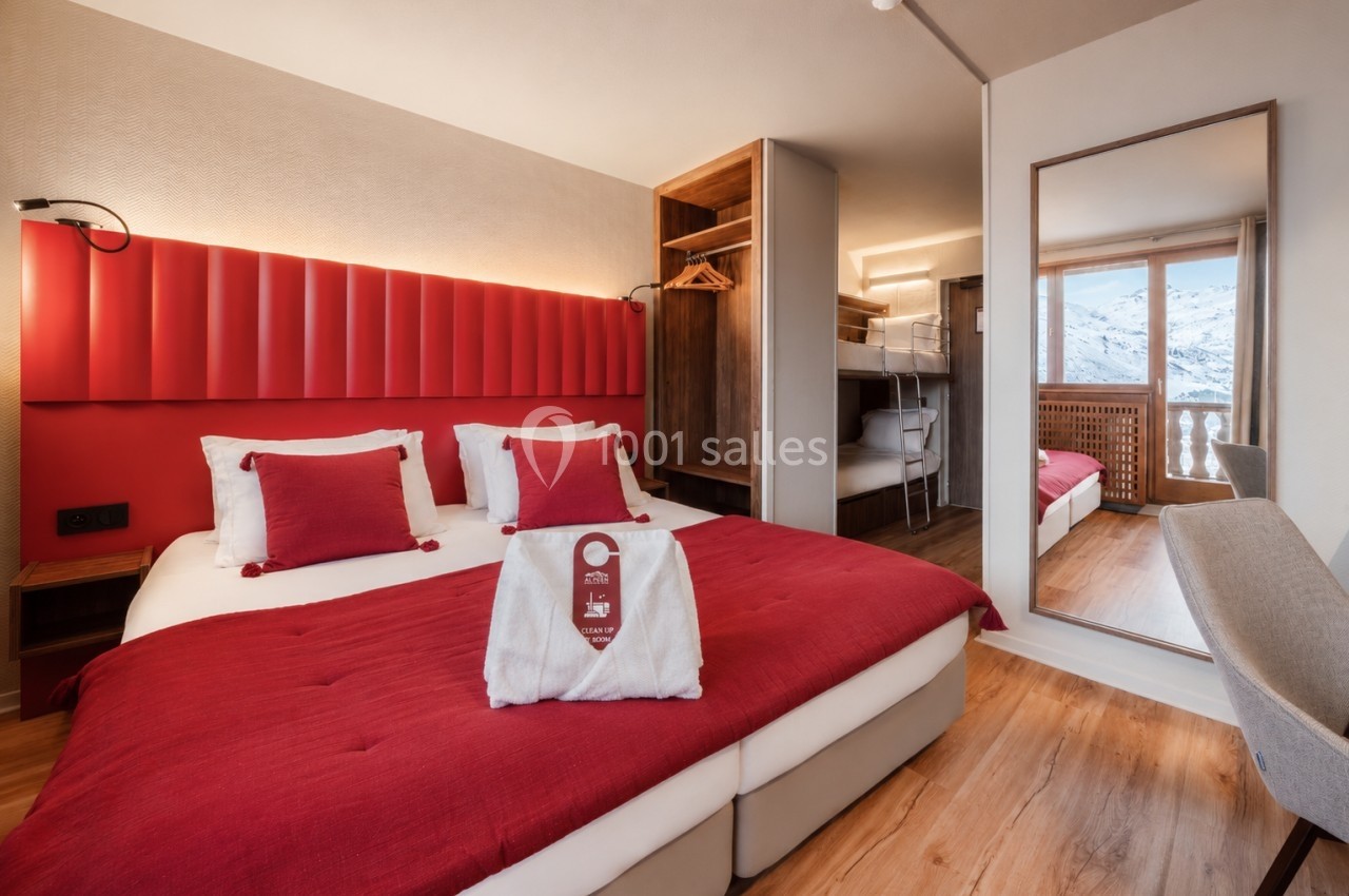 Chambre d'hôtel avec lit double, lits superposés, miroir, et vue sur des montagnes enneigées depuis le balcon.