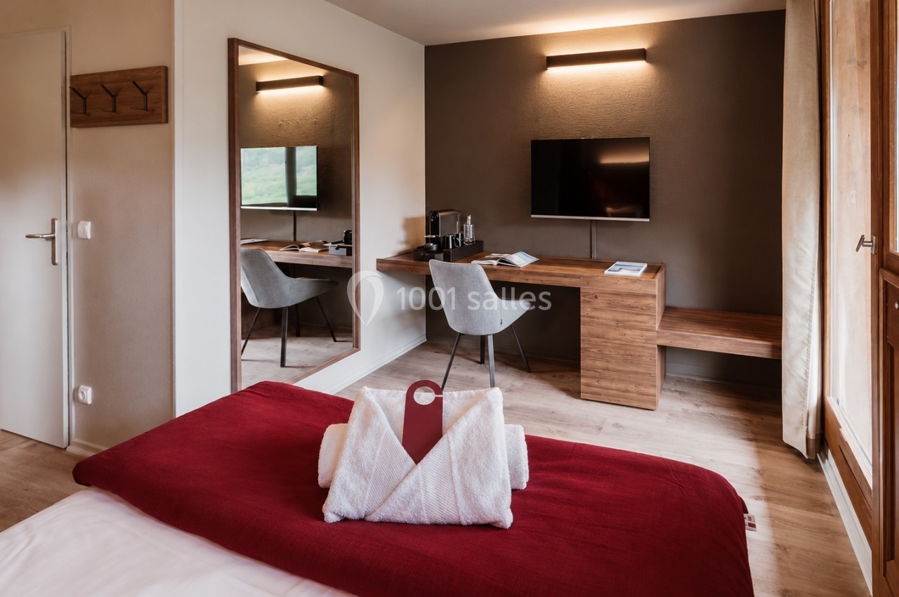 Chambre d'hôtel avec lit couvert d'un plaid rouge, bureau en bois, chaise grise et télévision murale.