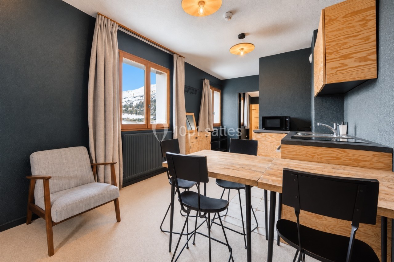 Salle à manger avec table en bois, chaises noires, coin cuisine équipé et fenêtre offrant une vue sur la montagne enneigée.