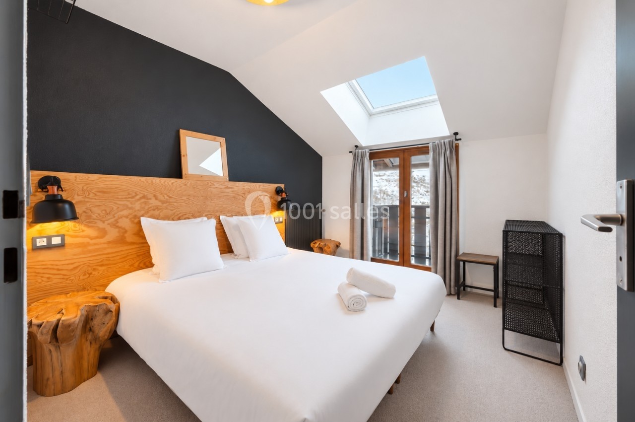 Chambre lumineuse avec lit double, murs blancs, tête de lit en bois, fenêtre avec rideaux et vue sur l'extérieur.