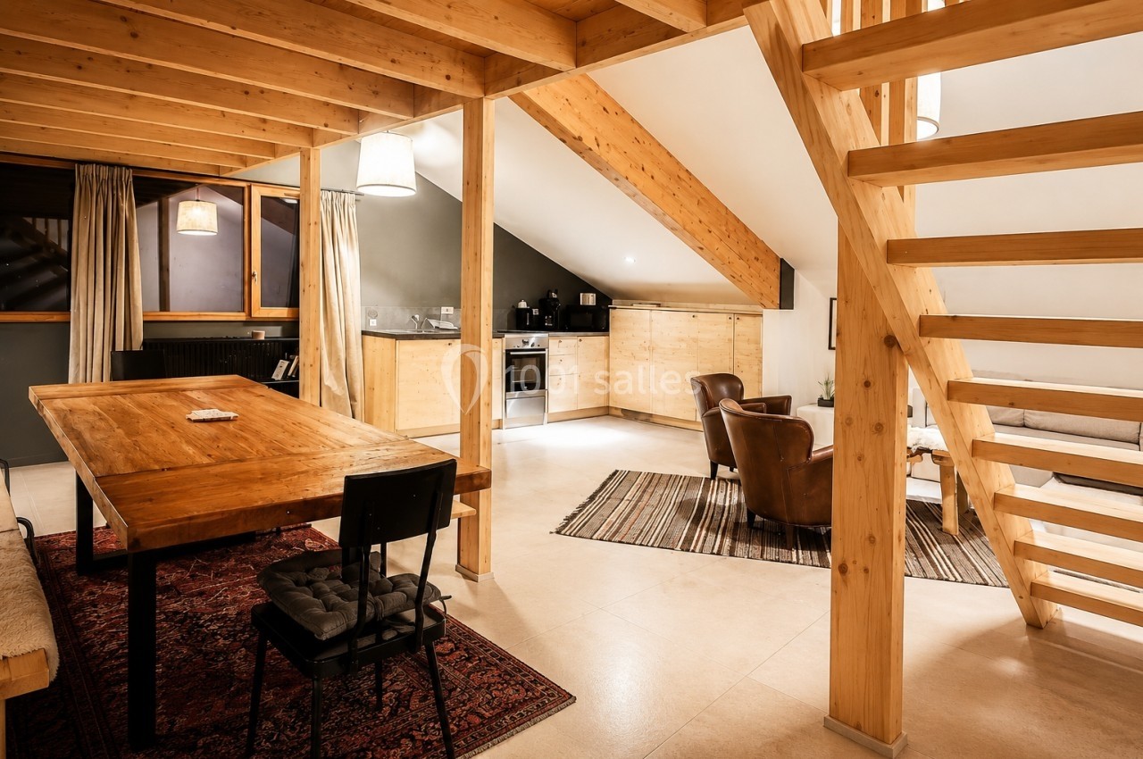 Intérieur d'un salon chaleureux avec poutres apparentes, table en bois, cuisine ouverte et escalier en bois.