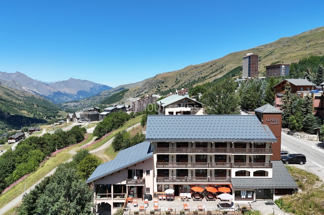 Vue aérienne d'un hôtel entouré de montagnes, avec des bâtiments voisins et un ciel dégagé en arrière-plan.