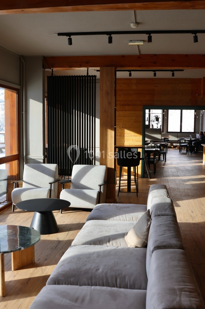 Salon moderne avec canapé gris, fauteuils et tables basses, éclairé par la lumière naturelle provenant de grandes fenêtres.