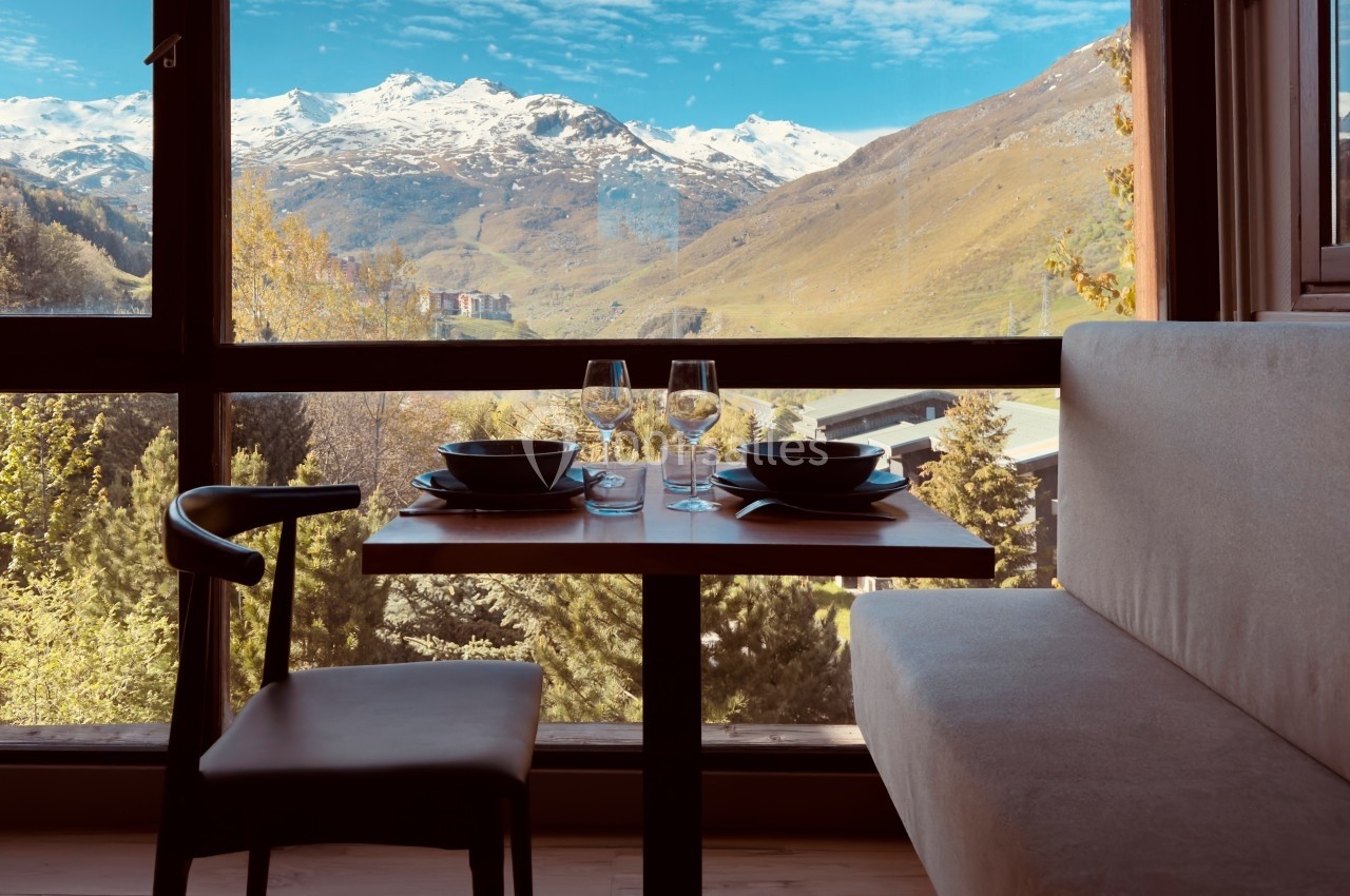 Table dressée pour deux avec vue sur des montagnes enneigées et une vallée verdoyante à travers une grande baie vitrée.