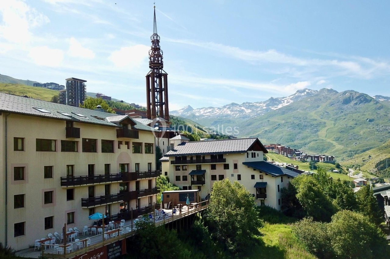 Vue d'un complexe hôtelier alpin avec une tour distinctive, entouré de montagnes et de verdure sous un ciel dégagé.