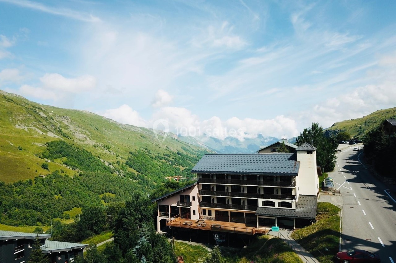 Bâtiment de montagne avec balcons en bois, entouré de collines verdoyantes sous un ciel partiellement nuageux.