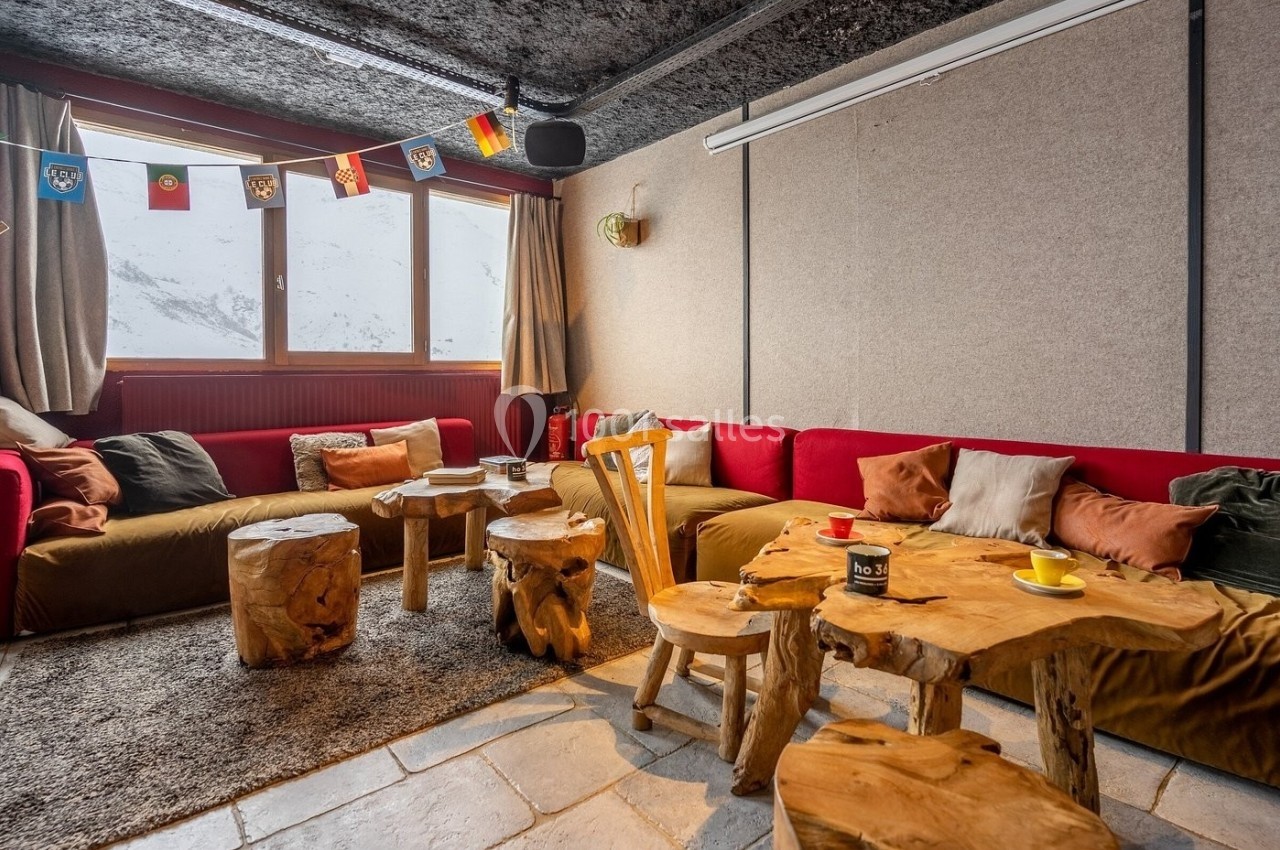 Salon chaleureux avec canapés, tables en bois brut, tapis et vue sur des montagnes enneigées par les fenêtres.