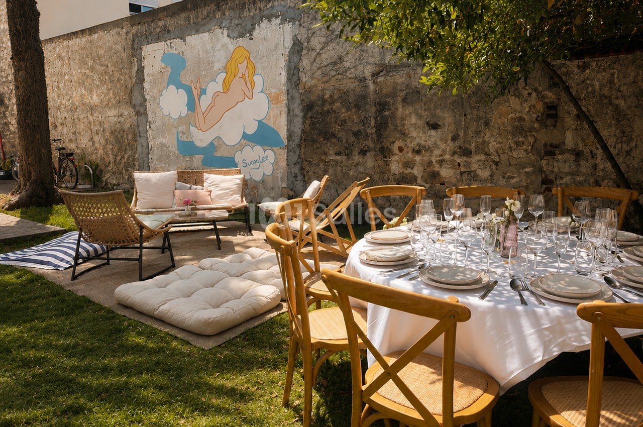 Table dressée en terrasse avec chaises bois, verres et couverts, canapé et murs en pierre brute au fond.