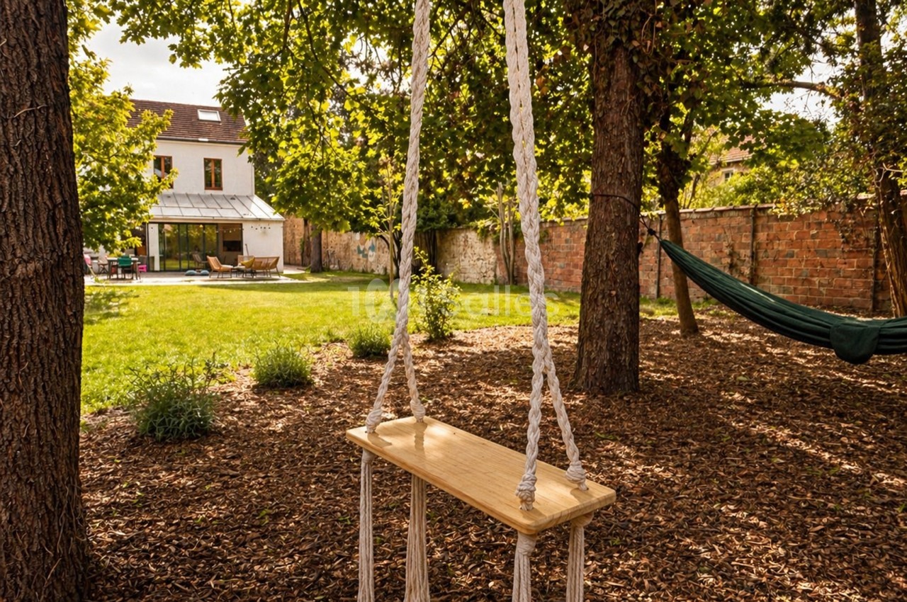Jardin ombragé avec balançoire en bois suspendue aux grands arbres, maison et terrasse en arrière-plan.