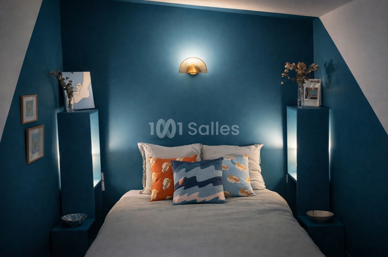 Chambre aux murs bleu pétrole, lit avec coussins colorés, deux petites tables de nuit, applique murale dorée et tableaux.