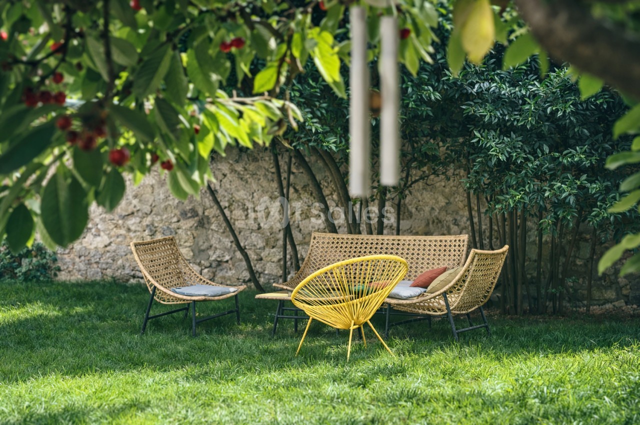 Jardin avec trois fauteuils en rotin tressé - deux beige, un jaune - sur herbe verte, mur de pierre et végétation.