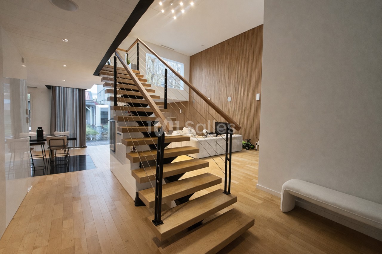 Escalier central en bois avec rampe métallique noire dans un espace intérieur aux murs blanc et bois clair.