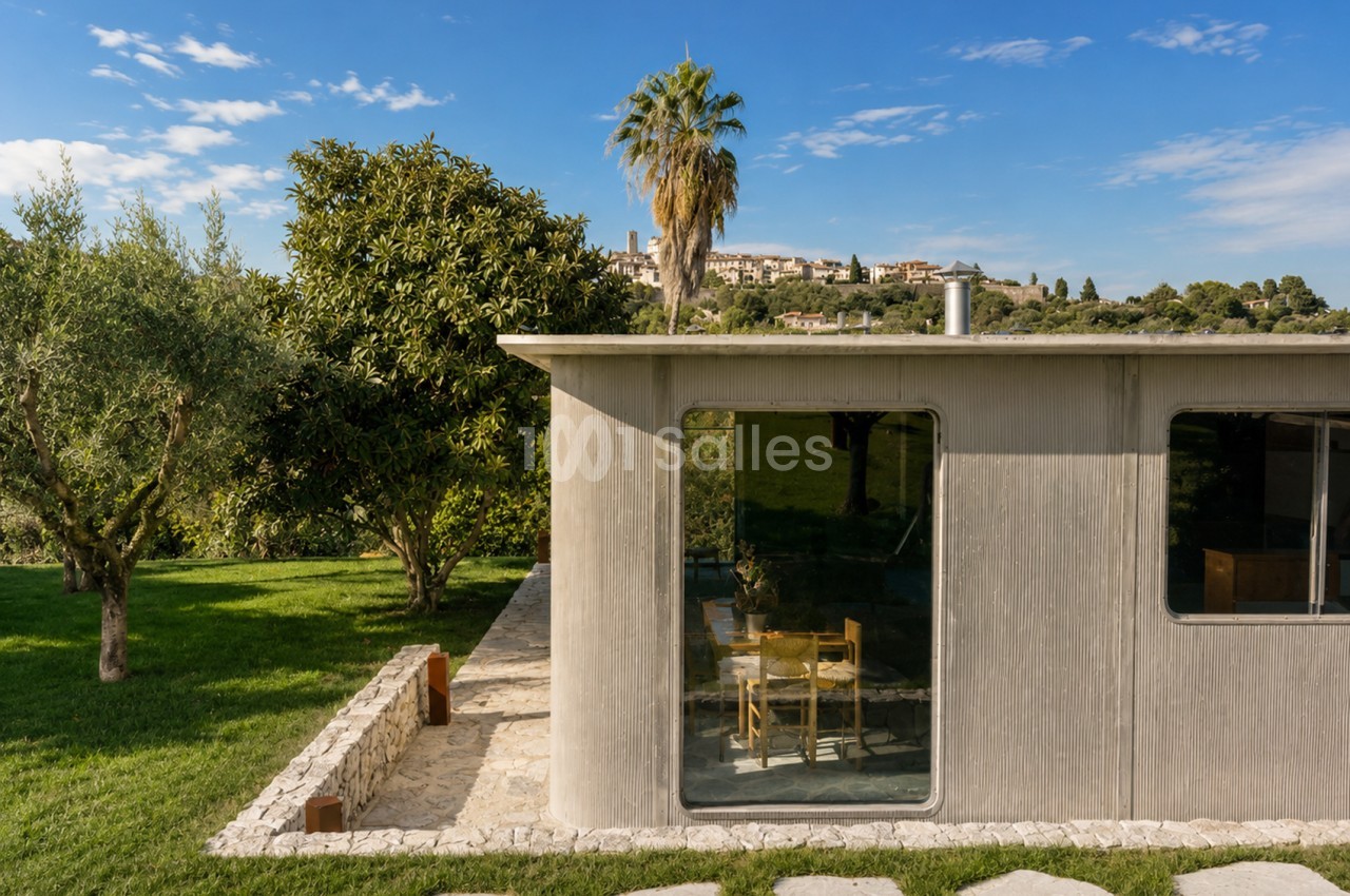 Bâtiment moderne avec façade ondulée beige, baies vitrées, entouré d'oliviers et pelouse, vue sur village provençal en arrière-plan.