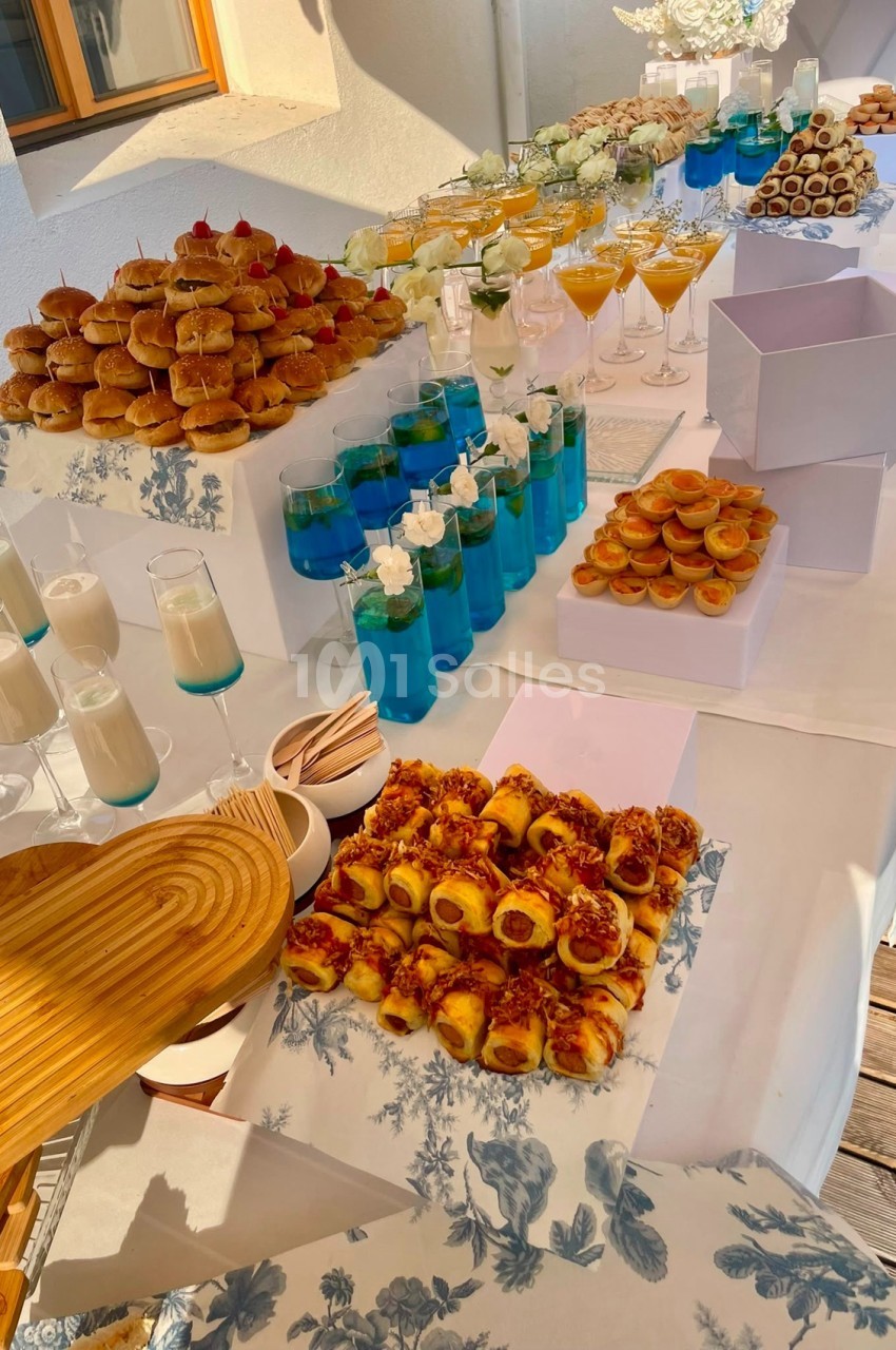 Table dressée en extérieur avec canapés variés, verres de cocktails bleus et blancs, fleurs blanches, sous lumière naturelle.