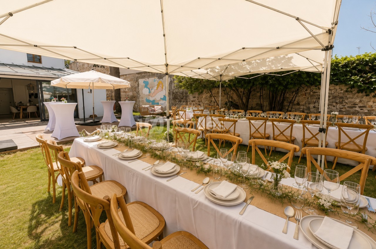 Salle extérieure dressée sous toile blanche avec tables longues nappe blanche, chaises bois, centre de table floral et couverts - jardin avec murs pierre et végétation.