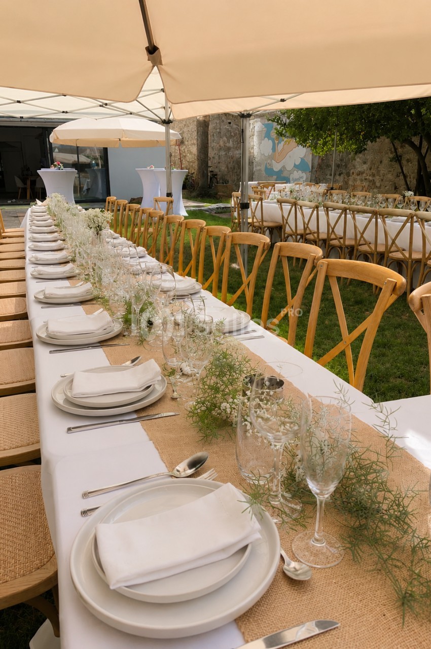 Table dressée en longueur sous tonnelle beige, chaises en bois blond, couverts blancs et verres avec branchages, terrasse et murs en pierre.