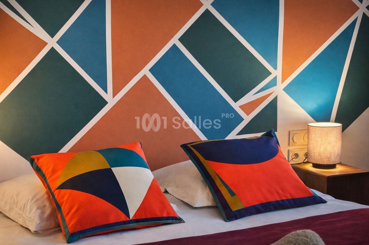 Chambre avec deux coussins géométriques colorés sur lit blanc et mur peint de losanges orange, bleu et vert.