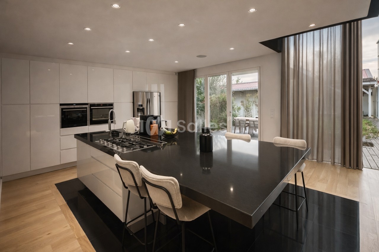 Cuisine moderne avec îlot central noir, deux chaises hautes, électroménagers intégrés et baies vitrées ouvrant sur terrasse.