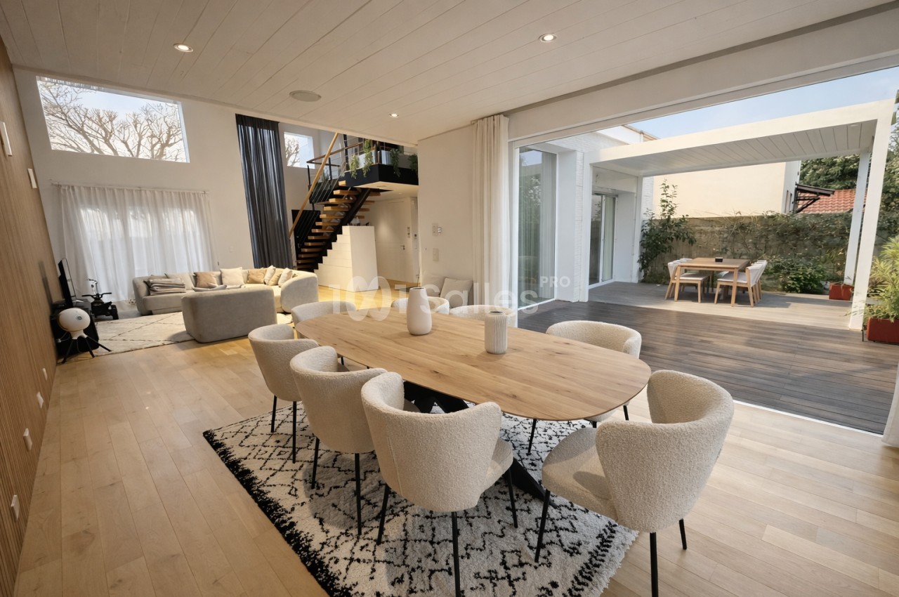 Intérieur contemporain avec salle à manger au premier plan, table en bois, chaises beiges, salon arrière, escalier noir, baies vitrées ouvertes sur terrasse.