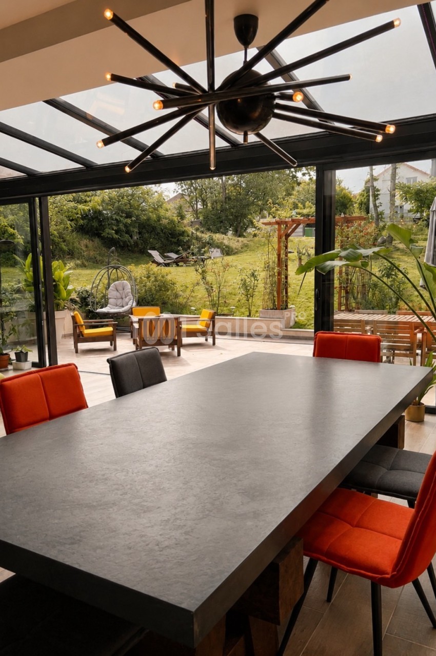 Salle couverte avec table grise, chaises oranges et rouges, lustre design noir, vue sur jardin arborisé.