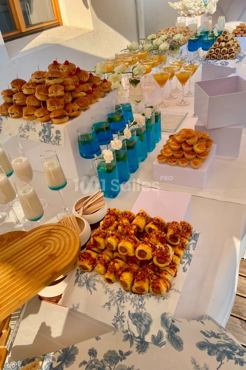 Table dressée avec canapés garnis, verres de cocktails bleus et jaunes, fleurs blanches, en terrasse ensoleillée.