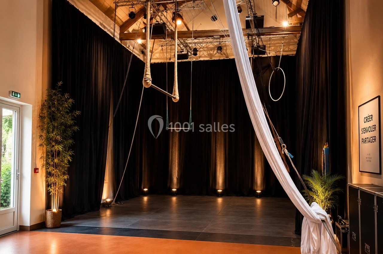 Salle de spectacle avec scène vide, rideaux noirs, équipements aériens suspendus et éclairage tamisé.