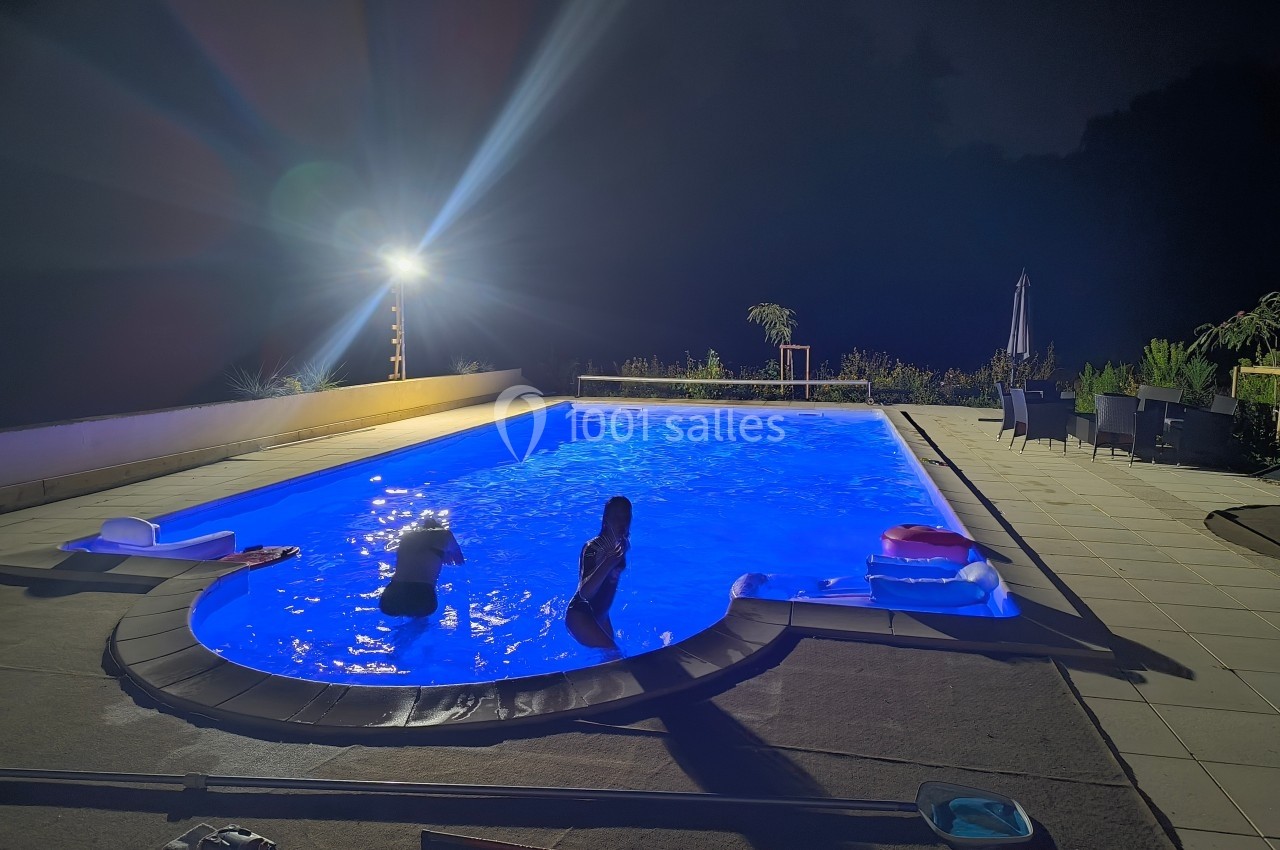Piscine éclairée de nuit avec deux personnes dans l'eau, entourée de chaises et de végétation.