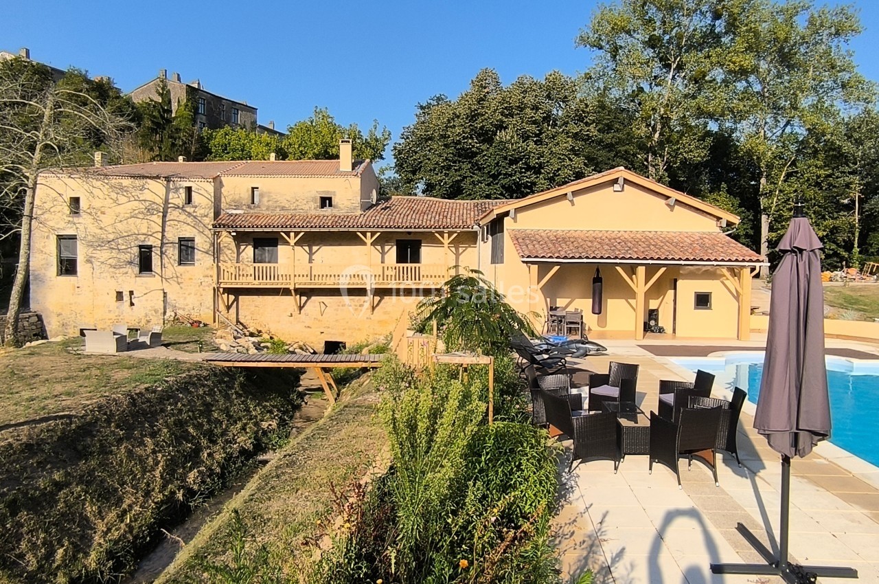 Maison en pierre avec terrasse, jardin, piscine et mobilier d'extérieur, entourée de verdure sous un ciel dégagé.